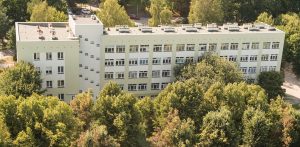 Z lotu ptaka widać Szpital Dziecięcy Polanki im. Macieja Płażyńskiego w Gdańsku. To duży, kilkupiętrowy budynek o jasnozielonej elewacji z licznymi oknami z płaskim dachem, na którym widać różne elementy wentylacyjne i techniczne. Szpital otoczony jest gęstym lasem liściastym. W tle, za drzewami, widać fragmenty innych budynków i parking