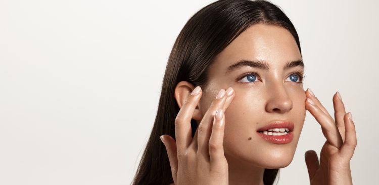 Skin care and women cosmetology. Woman rubbing cream, skincare beauty product on her face, looking away while filling under eye skin with hydrating and nourishing cosmetic products, white background.