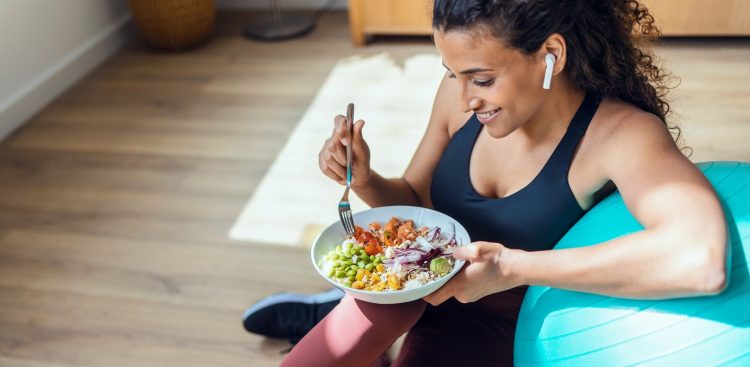 Shot of sporty young woman eating healthy while listening to music sitting on the floor at home.