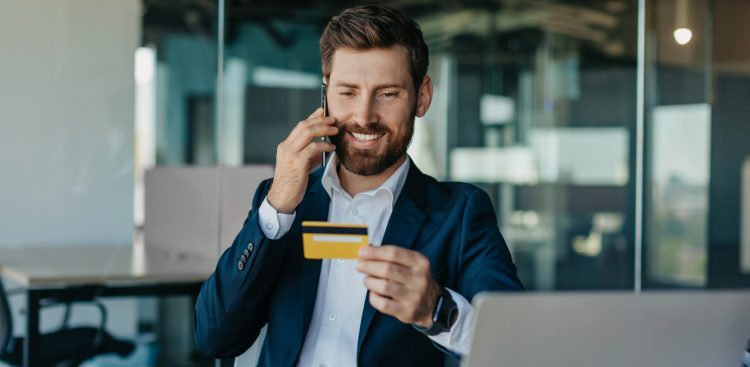 Mobile payments. Happy male worker holding credit card after online purchase and talking on mobile phone with customer service, sitting at workplace in office, copy space