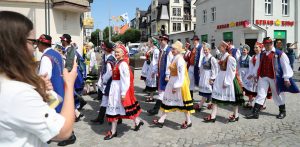 Przemarsz w czasie zjazdu Kaszubów. Grupa osób ubranych w tradycyjne, kolorowe stroje kaszubskie maszeruje podczas parady po brukowanej ulicy w słoneczny dzień. Kobiety mają na sobie białe, haftowane bluzki, ciemne gorsety, barwne spódnice (w odcieniach czerwieni, żółci i zieleni) oraz ozdobne wianki lub tradycyjne czepce. Mężczyźni noszą niebieskie, wyszywane kamizelki, białe koszule, jasne spodnie i ciemne kapelusze z czerwonymi elementami. W tle widoczna jest miejska architektura z szyldem lokalu „Kebab King”. Na pierwszym planie po lewej stronie kobieta trzyma smartfon, robiąc zdjęcie maszerującemu korowodowi