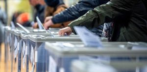 Grupa ludzi, którzy wrzucają głosy do urn wyborczych. Widoczne są dłonie wrzucające karty do przezroczystych urn. W tle widać niewyraźne sylwetki innych osób.