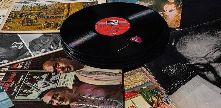 Artystyczna kompozycja ze stosem płyt winylowych i okładkami albumów z muzyką. Na wierzchniej płycie leży mały różowy kwiat