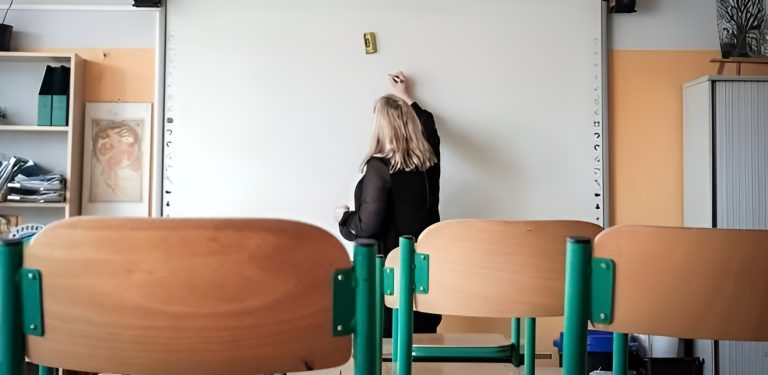 Nauczycielka w sali lekcyjnej przy białej tablicy, na pierwszym planie puste krzesła