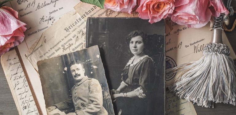 Kompozycja składająca się ze starych dokumentów (świadectw, metryk), dwóch wyblakłych czarno-białych fotografii (portret kobiety i mężczyzny z wąsami) oraz suszonych różowych róż i ozdobnego chwosta, symbolizująca pamięć i historię rodziny.