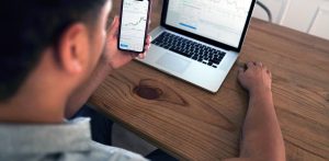 Mężczyzna spogląda na wykresy inwestycyjne w smartfonie, przed nim stoi laptop, na ekranie również wykresy