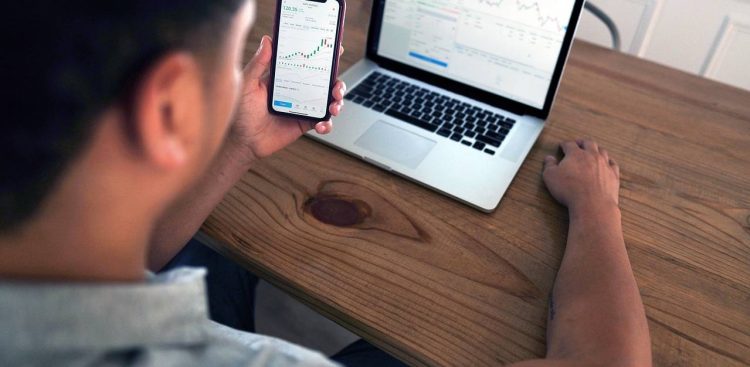 Mężczyzna spogląda na wykresy inwestycyjne w smartfonie, przed nim stoi laptop, na ekranie również wykresy