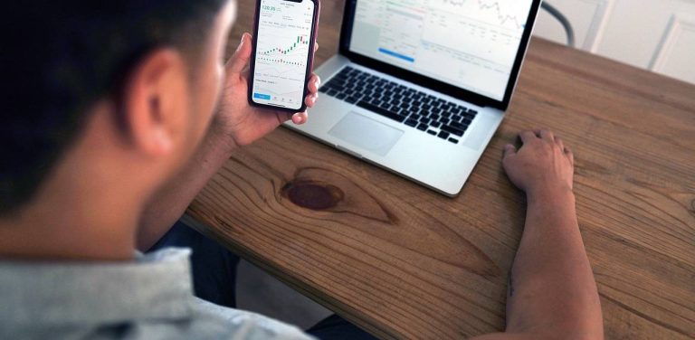 Mężczyzna spogląda na wykresy inwestycyjne w smartfonie, przed nim stoi laptop, na ekranie również wykresy