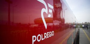 Zbliżenie na czerwony bok wagonu pociągu Polregio z wyraźnym białym logo przedstawiającym stylizowanego ptaka oraz napisem „POLREGIO”. W tle widać lekko rozmyty fragment peronu kolejowego i sylwetki ludzi