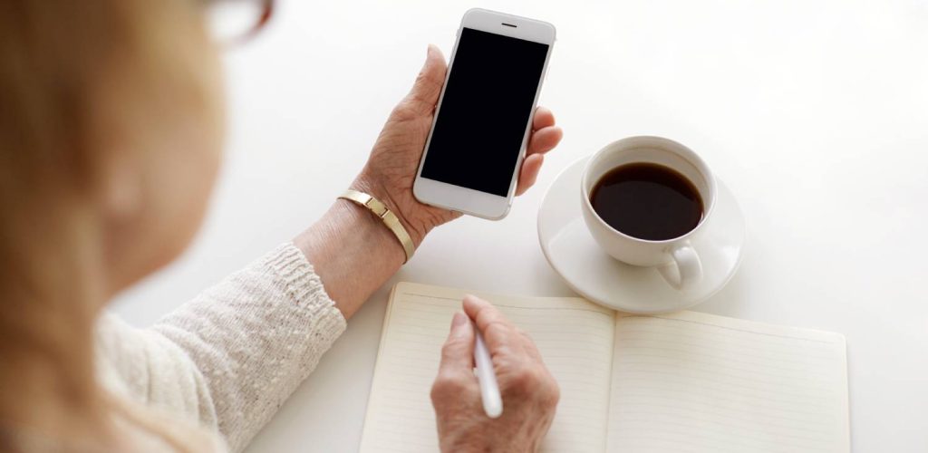 Kobieta trzyma telefon i zapisuje coś w notesie, przed nią stoi kawa