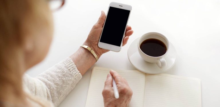 Kobieta trzyma telefon i zapisuje coś w notesie, przed nią stoi kawa