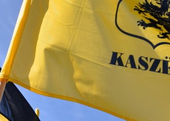 Dwie powiewające na tle nieba kaszubskie flagi. Po prawej stronie widoczna jest żółta flaga z czarnym gryfem i drukowanym napisem „KASZËBË”, natomiast po lewej stronie znajduje się fragment dwubarwnej, czarno-żółtej flagi zamocowanej na drewnianym drzewcu