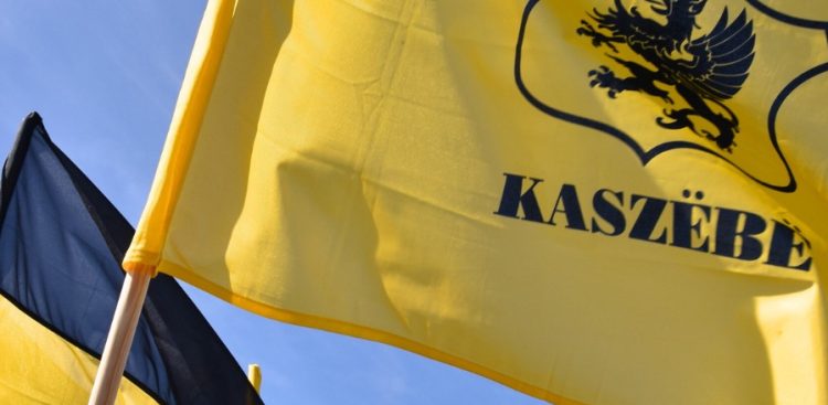 Dwie powiewające na tle nieba kaszubskie flagi. Po prawej stronie widoczna jest żółta flaga z czarnym gryfem i drukowanym napisem „KASZËBË”, natomiast po lewej stronie znajduje się fragment dwubarwnej, czarno-żółtej flagi zamocowanej na drewnianym drzewcu