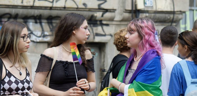 Zgromadzenie młodzieży, dwie dziewczyny patrzą na siebie, jedna trzyma tęczową chorągiewkę, druga jest okryta flagą LGBT. W tle ściana kamienicy pokryta napisami