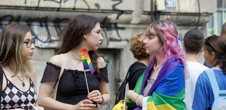 Zgromadzenie młodzieży, dwie dziewczyny patrzą na siebie, jedna trzyma tęczową chorągiewkę, druga jest okryta flagą LGBT. W tle ściana kamienicy pokryta napisami