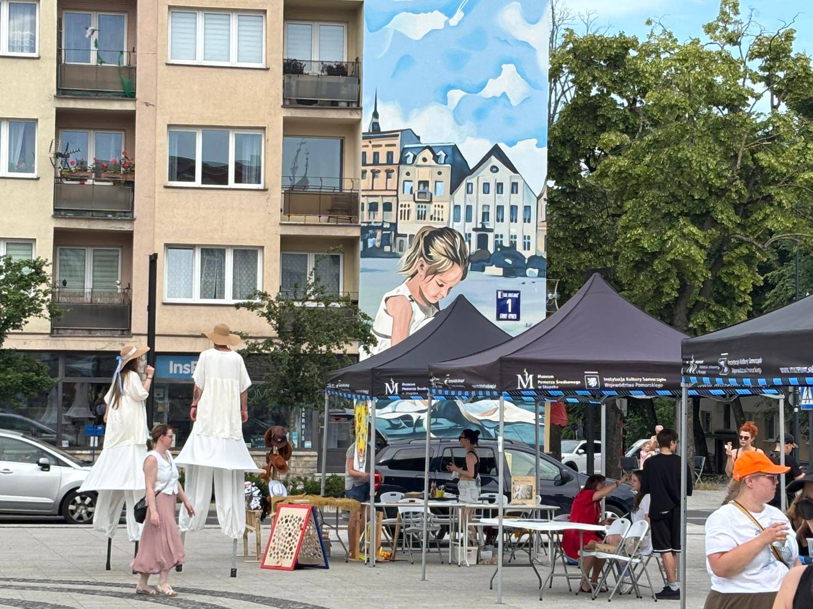 Szczudlarze i namioty warsztatowe Muzeum Pomorza Środkowego podczas festiwalu SALWA na Starym Rynku w Słupsku. W tle, na ścianie budynku, widnieje charakterystyczny słupski mural przedstawiający dziewczynkę na tle starówki 