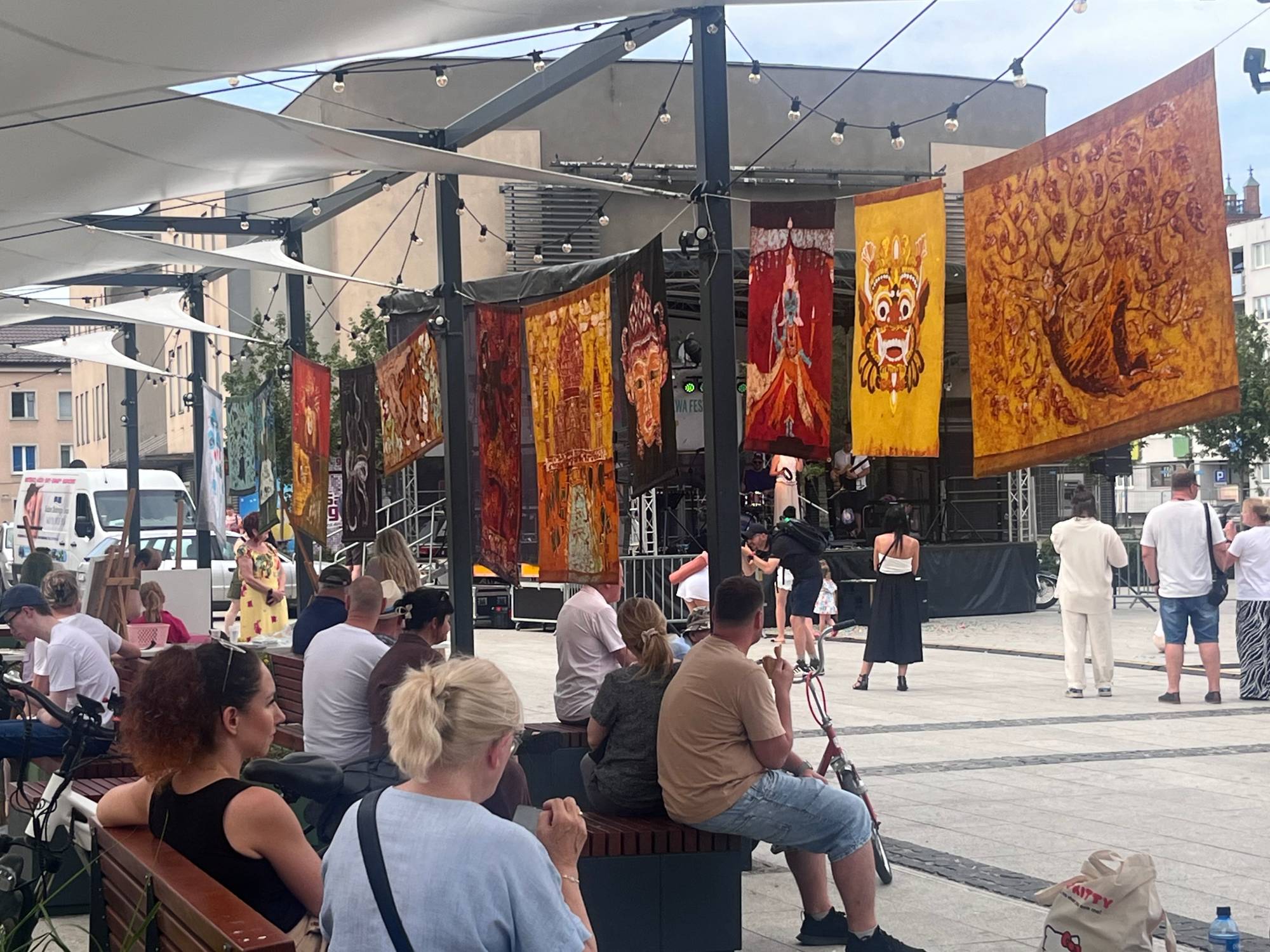 Widzowie siedzący na ławkach i oglądający koncert na festiwalu SALWA w Słupsku. Nad ich głowami wiszą kolorowe, ozdobne tkaniny o artystycznych wzorach, a w tle widać plenerową scenę muzyczną