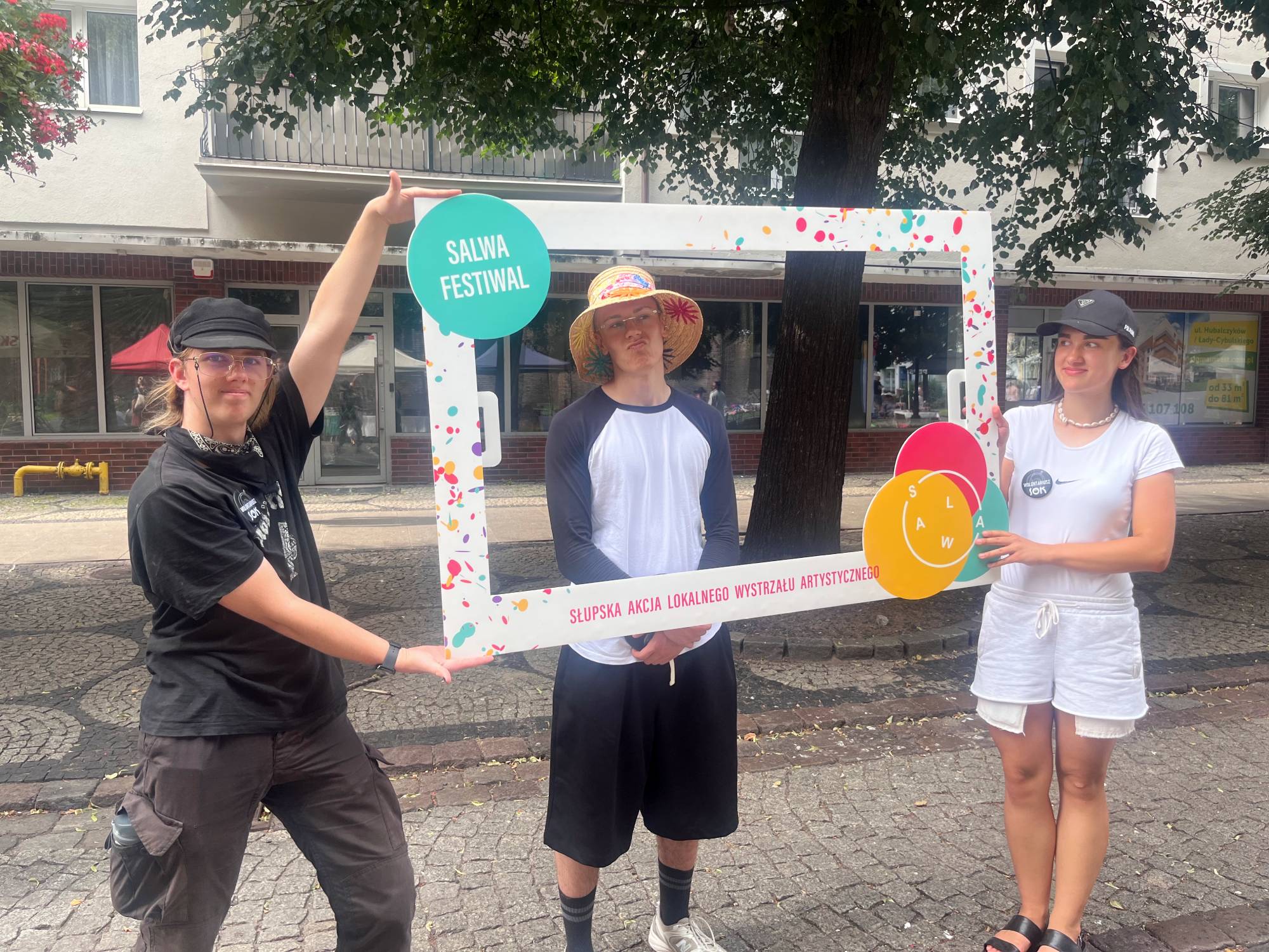 Troje młodych ludzi pozuje na placu podczas festiwalu SALWA w Słupsku. Dwie osoby po bokach trzymają dużą, białą ramkę fotograficzną ozdobioną kolorowym motywem konfetti oraz napisami „SALWA FESTIWAL” i „Słupska Akcja Lokalnego Wystrzału Artystycznego”. Chłopak pośrodku, w słomkowym kapeluszu, stoi wewnątrz ramki i patrzy w obiektyw. Osoby trzymające ramkę mają przypięte do koszulek okrągłe plakietki z napisem „Wolontariusz SOK”
