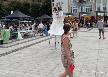 Szczudlarze w jasnych strojach i słomkowych kapeluszach na Starym Rynku w Słupsku podczas festiwalu SALWA, w otoczeniu spacerujących mieszkańców i namiotów wystawienniczych