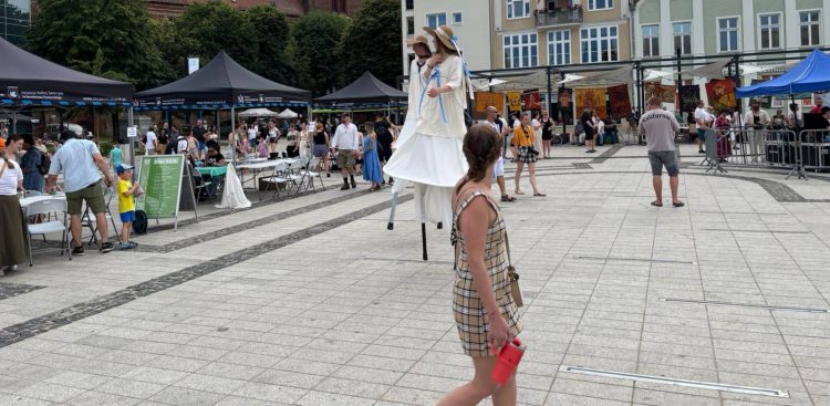 Szczudlarze w jasnych strojach i słomkowych kapeluszach na Starym Rynku w Słupsku podczas festiwalu SALWA, w otoczeniu spacerujących mieszkańców i namiotów wystawienniczych
