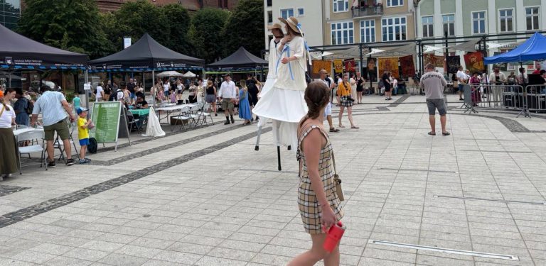Szczudlarze w jasnych strojach i słomkowych kapeluszach na Starym Rynku w Słupsku podczas festiwalu SALWA, w otoczeniu spacerujących mieszkańców i namiotów wystawienniczych