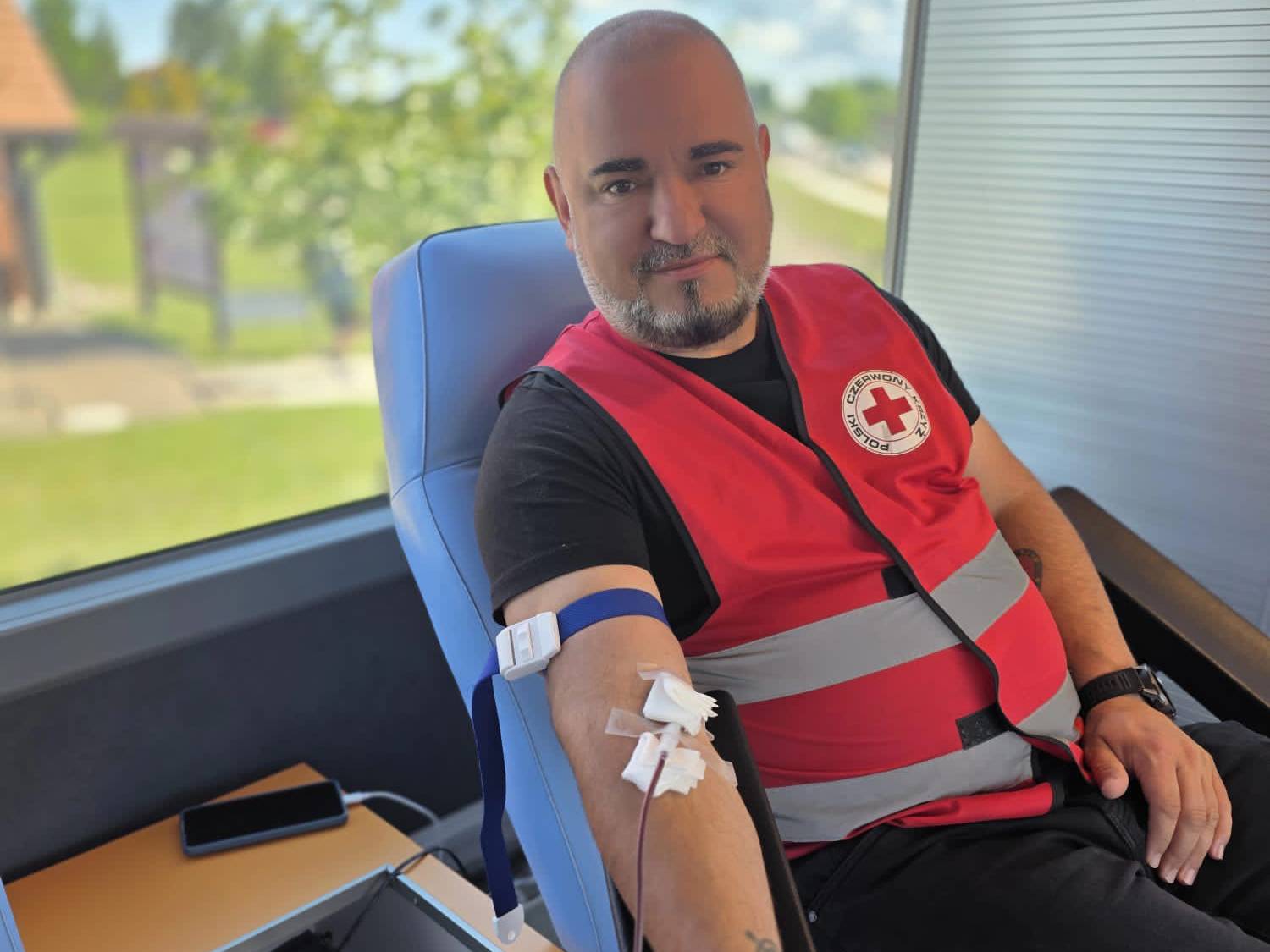 Mężczyzna w czerwonej kamizelce z napisem "Czerwony Krzyż Polski", siedzący na fotelu i oddający krew. Na jego ramieniu widoczna jest igła i rurka do pobierania krwi.