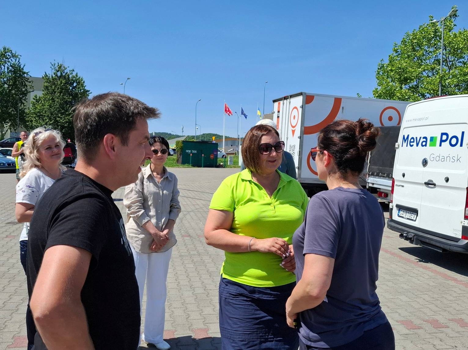 Prezydent Gdańska Aleksandra Dulkiewicz, widoczna od tyłu w ciemnoszarej koszulce i z włosami upiętymi w kok, rozmawia na wybrukowanym placu z wolontariuszką ubraną w jaskrawozieloną koszulkę polo i okulary przeciwsłoneczne. Po lewej stronie rozmowie przygląda się mężczyzna w czarnym t-shircie oraz dwie inne kobiety stojące nieco dalej. Po prawej stronie zaparkowany jest biały furgon dostawczy z napisem „Meva-Pol Gdańsk”, a za nim większa ciężarówka transportowa. W tle, pod bezchmurnym, niebieskim niebem, widoczne są maszty z flagami oraz zielone drzewa. Prezydent żegna wolontariuszy fundacji Zupa na Monciaku, którzy wyjeżdżają z 40. konwojem z pomocą na Ukrainę