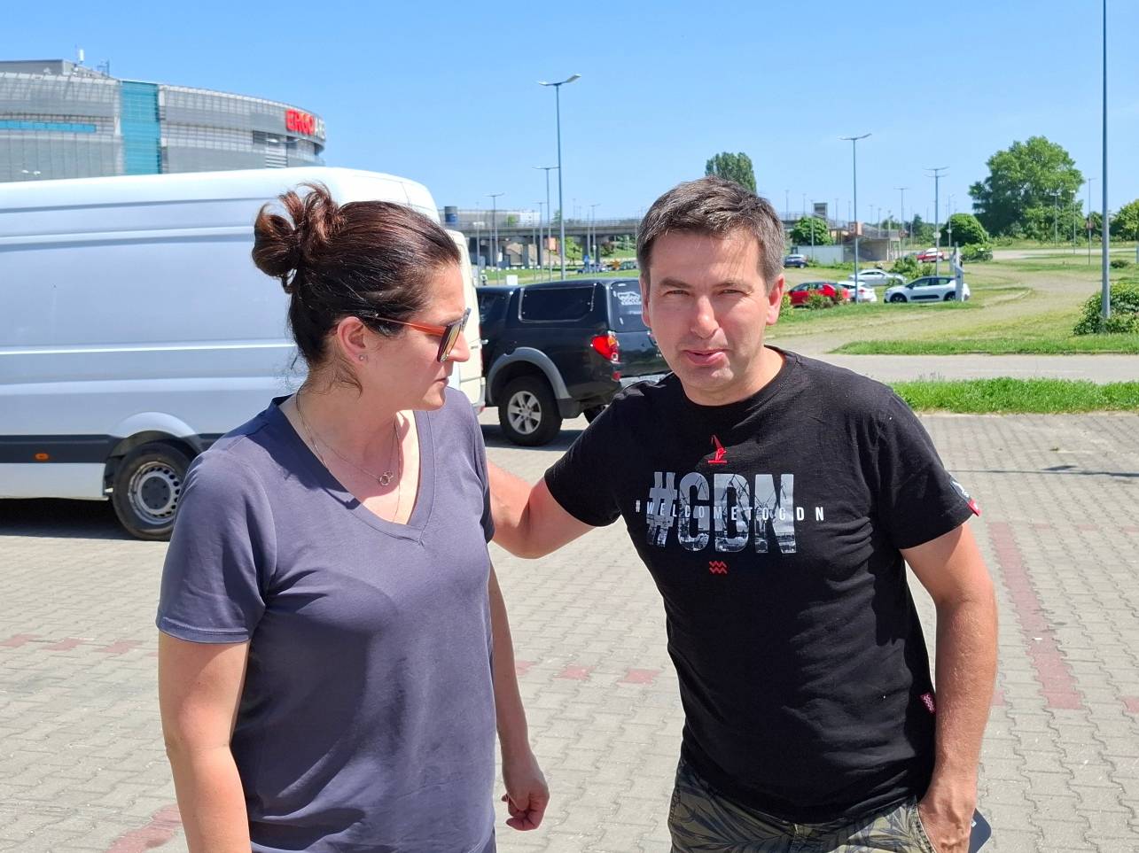 Zdjęcie przedstawia prezydent Gdańska Aleksandrę Dulkiewicz oraz mężczyznę w czarnym T-shircie z napisem „#GDN”. Oboje stoją obok siebie na brukowanym parkingu w słoneczny dzień. Kobieta ma spięte ciemne włosy, okulary i ciemnoszarą koszulkę. W tle, za ich plecami, widoczne są zaparkowane samochody (w tym biały dostawczy van) oraz w oddali zarys hali Ergo Arena na granicy Gdańska i Sopotu