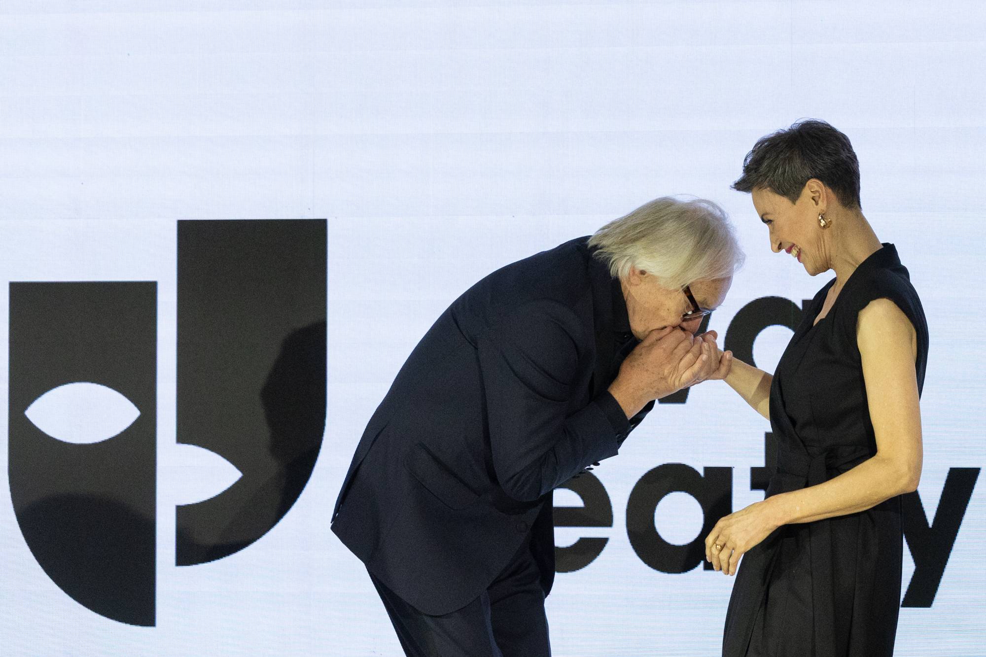Andrzej Seweryn i Danuta Stenka na scenie podczas tej gali festiwalu „Dwa Teatry”. Andrzej Seweryn, ubrany w ciemny garnitur i okulary, pochyla się w pełnym szacunku geście, całując dłoń aktorki. Danuta Stenka, stojąca po prawej stronie kadru w czarnej kreacji bez rękawów, patrzy na niego z góry z ciepłym, serdecznym uśmiechem. W tle znajduje się duży, jasny ekran z czarnym logotypem festiwalu – stylizowaną maską teatralną