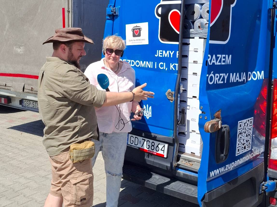 Dziennikarka Anna Rębas w okularach przeciwsłonecznych trzyma niebieski mikrofon Radia Gdańsk, nagrywając wypowiedź mężczyzny ubranego w oliwkową koszulę, krótkie spodenki i brązowy kapelusz turystyczny. Stoją na brukowanym placu przy otwartym tyle niebieskiego furgonu transportowego z logo fundacji „Zupa na Monciaku”. Wewnątrz pojazdu widać ułożone w stosy kartony i paczki z pomocą humanitarną. Na karoserii furgonu, obok tablicy rejestracyjnej z Gdańska, naklejona jest naklejka z herbem Ukrainy (Tryzubem) oraz widoczny jest adres strony internetowej fundacji
