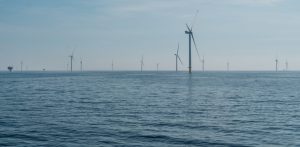 Morska farma wiatrowa Baltic Power. Szerokie ujęcie przedstawiające morską farmę wiatrową na otwartym morzu. Na pierwszym planie widoczna jest ciemnoniebieska, lekko pofalowana powierzchnia wody. W oddali, na linii horyzontu i przed nią, wznosi się kilkanaście białych turbin wiatrowych. Jedna z nich, usytuowana po prawej stronie kadru, znajduje się bliżej i jest wyraźniejsza, podczas gdy pozostałe są rozproszone w tle i maleją wraz z odległością. Nad horyzontem rozciąga się jasnobłękitne, lekko zamglone niebo. Po lewej stronie, na samym skraju kadru w oddali, majaczy zarys małej platformy morskiej