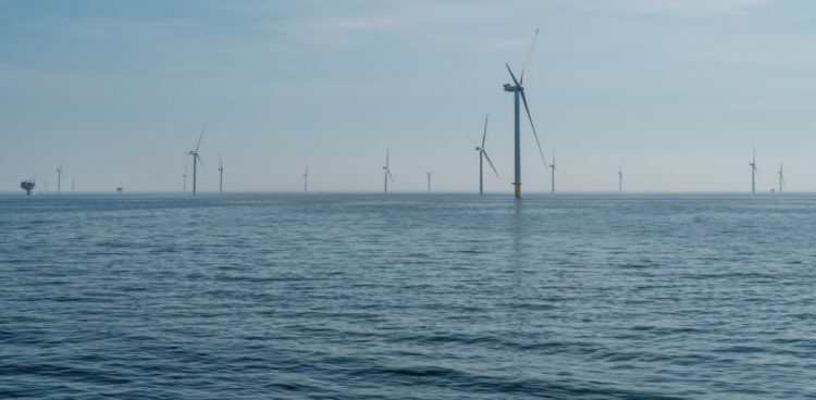 Morska farma wiatrowa Baltic Power. Szerokie ujęcie przedstawiające morską farmę wiatrową na otwartym morzu. Na pierwszym planie widoczna jest ciemnoniebieska, lekko pofalowana powierzchnia wody. W oddali, na linii horyzontu i przed nią, wznosi się kilkanaście białych turbin wiatrowych. Jedna z nich, usytuowana po prawej stronie kadru, znajduje się bliżej i jest wyraźniejsza, podczas gdy pozostałe są rozproszone w tle i maleją wraz z odległością. Nad horyzontem rozciąga się jasnobłękitne, lekko zamglone niebo. Po lewej stronie, na samym skraju kadru w oddali, majaczy zarys małej platformy morskiej