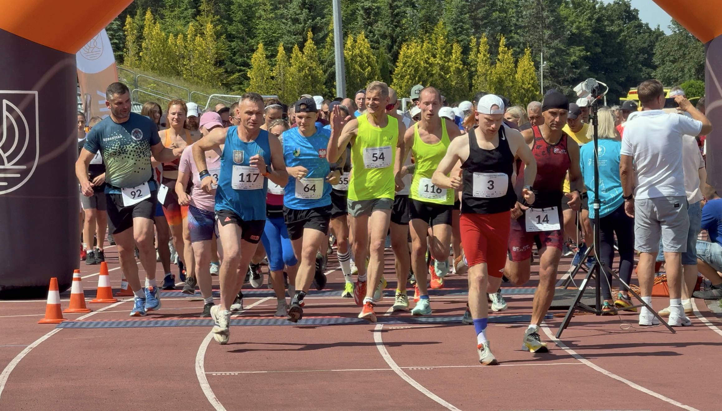 Widok od przodu na grupę biegaczy startujących w biegu charytatywnym na czerwonej bieżni lekkoatletycznej. Zawodnicy w kolorowych strojach sportowych z przypiętymi numerami startowymi biegną w stronę aparatu spod dmuchanej bramy w słoneczny dzień. Impreza to "Bieg z Nadzieją" w Słupsku
