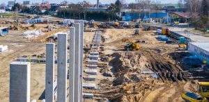Budowa zakładu firmy Iglotex w Skórczu (woj. pomorskie). Duży plac budowy widziany z góry. Na pierwszym planie widać rzędy pionowych betonowych słupów, które wyglądają na część nowo powstającej konstrukcji. Za nimi rozciąga się rozległy teren budowy z nierównym, ziemistym podłożem, na którym pracują koparki i inny ciężki sprzęt budowlany. Jasne niebo w słoneczny dzień