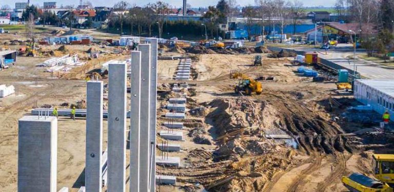 Budowa zakładu firmy Iglotex w Skórczu (woj. pomorskie). Duży plac budowy widziany z góry. Na pierwszym planie widać rzędy pionowych betonowych słupów, które wyglądają na część nowo powstającej konstrukcji. Za nimi rozciąga się rozległy teren budowy z nierównym, ziemistym podłożem, na którym pracują koparki i inny ciężki sprzęt budowlany. Jasne niebo w słoneczny dzień