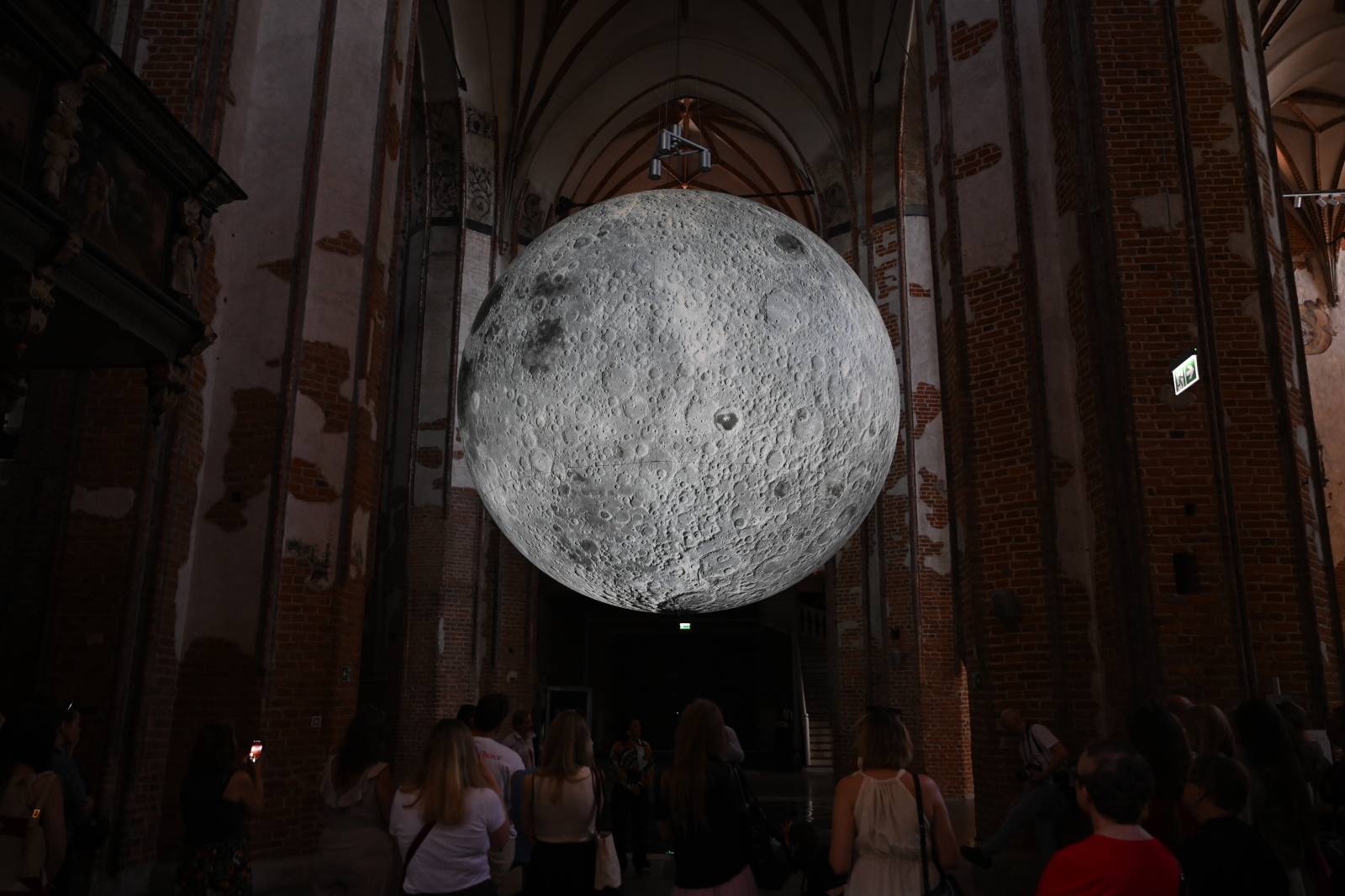 Instalacja „Museum of the Moon" w Centrum św. Jana w Gdańsku. Duża świecąca replika Księżyca zawieszona wysoko pod sklepieniem mrocznego kościoła o ceglanych filarach. Na dole widoczne są sylwetki ludzi przyglądających się instalacji 