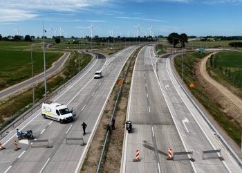 Widok z góry na zbliżenie tymczasowej blokady na nowo wybudowanym odcinku drogi ekspresowej S6. Na obu jezdniach ustawiono metalowe zapory oraz pionowe, biało-czerwone pachołki ostrzegawcze uniemożliwiające ruch. Na lewym pasie stoi biały furgon techniczny z otwartymi drzwiami kabiny, policyjny motocykl oraz jedna osoba w ciemnym ubiorze. Na prawej jezdni, tuż przy barierze energochłonnej oddzielającej kierunki ruchu, zaparkowany jest drugi policyjny motocykl, a obok niego stoją dwie osoby. W oddali, za blokadą, lewym pasem jedzie inny biały samochód dostawczy. Po obu stronach trasy rozciągają się zielone pola uprawne, a na horyzoncie widoczna jest farma wiatrowa z białymi wiatrakami pod błękitnym niebem