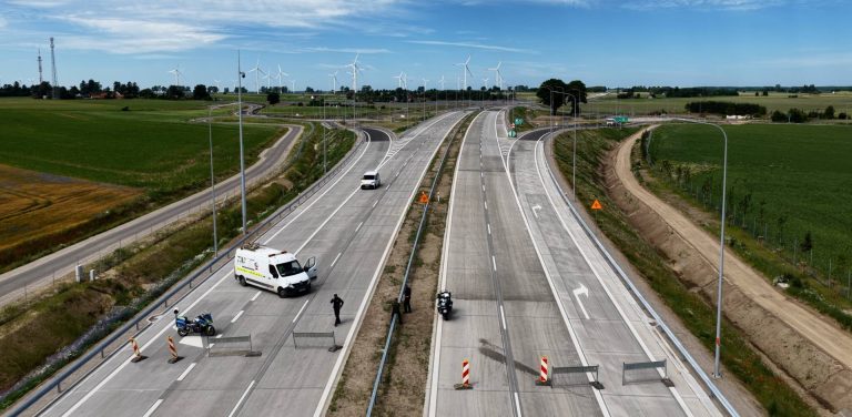 Widok z góry na zbliżenie tymczasowej blokady na nowo wybudowanym odcinku drogi ekspresowej S6. Na obu jezdniach ustawiono metalowe zapory oraz pionowe, biało-czerwone pachołki ostrzegawcze uniemożliwiające ruch. Na lewym pasie stoi biały furgon techniczny z otwartymi drzwiami kabiny, policyjny motocykl oraz jedna osoba w ciemnym ubiorze. Na prawej jezdni, tuż przy barierze energochłonnej oddzielającej kierunki ruchu, zaparkowany jest drugi policyjny motocykl, a obok niego stoją dwie osoby. W oddali, za blokadą, lewym pasem jedzie inny biały samochód dostawczy. Po obu stronach trasy rozciągają się zielone pola uprawne, a na horyzoncie widoczna jest farma wiatrowa z białymi wiatrakami pod błękitnym niebem