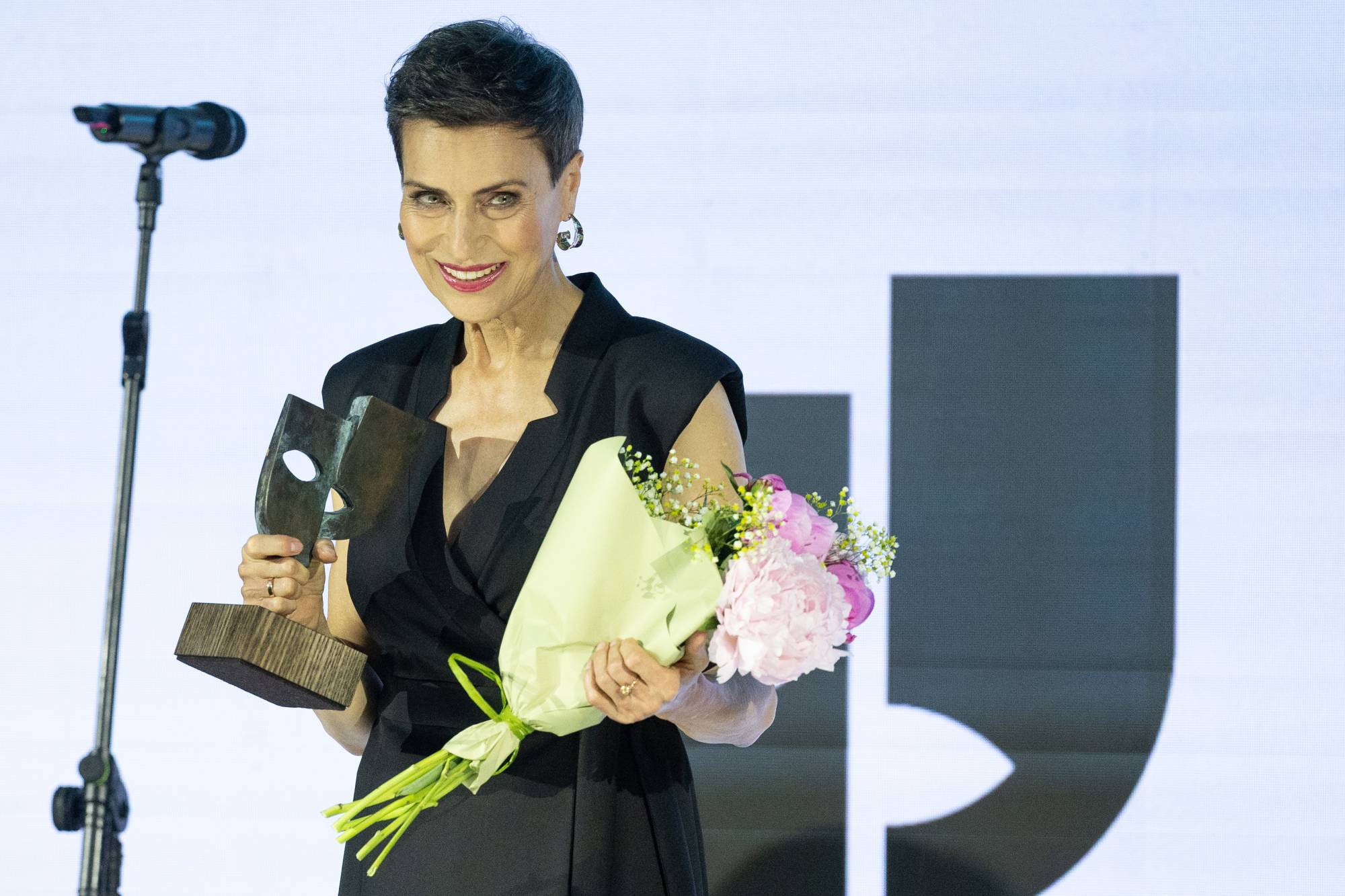 Zdjęcie przedstawia Danutę Stenkę stojącą na scenie podczas gali Festiwalu „Dwa Teatry” w Sopocie. Aktorka o krótkich, ciemnych włosach patrzy przed siebie z szerokim, radosnym uśmiechem. Jest ubrana w tę samą czarną, elegancką kreację bez rękawów. W prawej dłoni dumnie trzyma statuetkę Wielkiej Nagrody Festiwalu – ciemną rzeźbę umieszczoną na prostopadłościennym, drewnianym cokole[1][2]. W lewej dłoni trzyma bukiet różowych i białych piwonii owinięty w jasnozielony papier. Po lewej stronie kadru widoczny jest mikrofon na wysokim statywie. W tle znajduje się jasny, podświetlany ekran z fragmentem dużej, czarnej grafiki