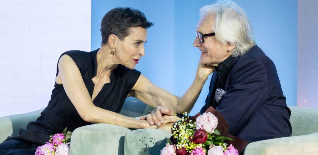Danuta Stenka i Andrzeja Seweryna siedzących obok siebie w jasnych fotelach podczas gali Festiwalu „Dwa Teatry”. Oboje są odwróceni do siebie profilami, patrzą sobie głęboko w oczy i ciepło się uśmiechają. Danuta Stenka, mająca krótkie, ciemne włosy i ubraną elegancką, czarną kreację bez rękawów, delikatnie trzyma lewą dłoń na policzku Andrzeja Seweryna, a prawą dłonią ściska jego rękę. Andrzej Seweryn – z półdługimi, siwymi włosami, w okularach w ciemnych oprawkach i ciemnej marynarce z poszetką – patrzy na nią z widocznym wzruszeniem. Na pierwszym planie, u dołu kadru, widoczne są bukiety różowych piwonii. Tło jest jednolite, w jasnoniebieskim odcieniu