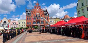 Dzień Strażaka i 160-lecie jednostki OSP w Chojnicach. Miejski rynek, w tle czerwony ratusz, odbywają się obchody Dnia Strażaka. Duża grupa strażaków w galowych mundurach stoi w szeregach. Na środku placu stoi podnośnik koszowy z biało-czerwoną flagą Polski, a przed nim odbywa się oficjalna część uroczystości. Po prawej stronie placu ustawione są czerwone namioty z napisem "PAŃSTWOWA STRAŻ POŻARNA", pod którymi również zgromadzili się uczestnicy. Błękitne niebo z białymi chmurami tworzy tło dla tego ważnego wydarzenia.