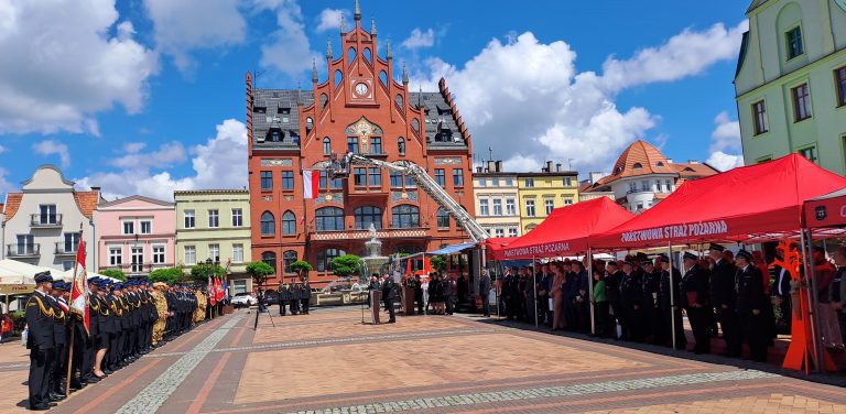 Dzień Strażaka i 160-lecie jednostki OSP w Chojnicach. Miejski rynek, w tle czerwony ratusz, odbywają się obchody Dnia Strażaka. Duża grupa strażaków w galowych mundurach stoi w szeregach. Na środku placu stoi podnośnik koszowy z biało-czerwoną flagą Polski, a przed nim odbywa się oficjalna część uroczystości. Po prawej stronie placu ustawione są czerwone namioty z napisem "PAŃSTWOWA STRAŻ POŻARNA", pod którymi również zgromadzili się uczestnicy. Błękitne niebo z białymi chmurami tworzy tło dla tego ważnego wydarzenia.
