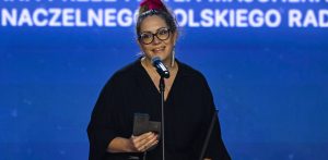 Zdjęcie przedstawia reżyserkę Magdalenę Małecką-Wippich stojącą na scenie przy mikrofonie podczas gali zamknięcia XXV edycji Festiwalu „Dwa Teatry – Sopot 2026”. Kobieta uśmiecha się, ma spięte na czubku głowy ciemne włosy z różową ozdobą, nosi okulary w ciemnych oprawkach, długie wiszące kolczyki oraz czarną kreację. W prawej dłoni trzyma ciemną, geometryczną statuetkę Grand Prix. W tle znajduje się jaskrawoniebieski ekran z fragmentami białych napisów