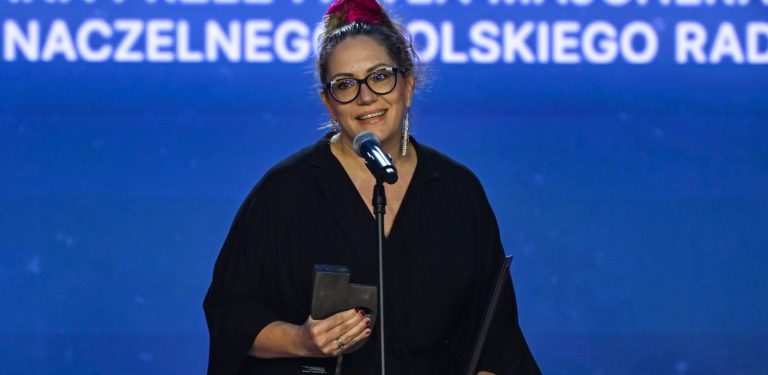 Zdjęcie przedstawia reżyserkę Magdalenę Małecką-Wippich stojącą na scenie przy mikrofonie podczas gali zamknięcia XXV edycji Festiwalu „Dwa Teatry – Sopot 2026”. Kobieta uśmiecha się, ma spięte na czubku głowy ciemne włosy z różową ozdobą, nosi okulary w ciemnych oprawkach, długie wiszące kolczyki oraz czarną kreację. W prawej dłoni trzyma ciemną, geometryczną statuetkę Grand Prix. W tle znajduje się jaskrawoniebieski ekran z fragmentami białych napisów