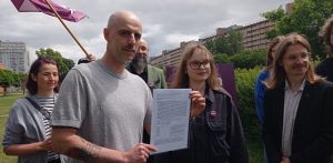 Mężczyzna z zarostem i tatuażami na przedramieniu trzyma dokument, obok niego stoi młoda kobieta w okularach. W tle widać grupę ludzi i fragment miasta, m.in. gdański "falowiec". Protest przeciwko zabudowie dzielnicy Przymorze