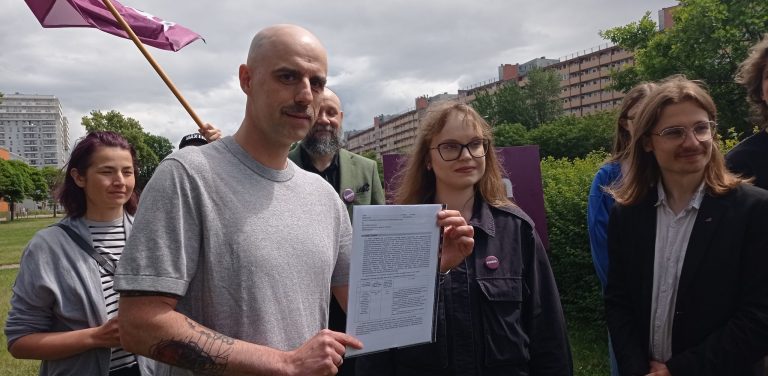 Mężczyzna z zarostem i tatuażami na przedramieniu trzyma dokument, obok niego stoi młoda kobieta w okularach. W tle widać grupę ludzi i fragment miasta, m.in. gdański "falowiec". Protest przeciwko zabudowie dzielnicy Przymorze