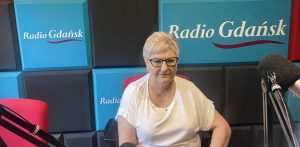Zdjęcie przedstawia Małgorzatę Żuchlińską, pielęgniarkę oddziałową Szpitalnego Oddziału Ratunkowego w Słupsku, siedzącą w studiu radiowym Radia Gdańsk. Ubrana jest w białą koszulkę, ma krótkie, jasne włosy i nosi okulary w ciemnych oprawkach. Jej wyraz twarzy jest poważny. Tłem jest ściana studia, wyłożona panelami akustycznymi w kolorach błękitnym i ciemnym szarym. Na panelach wyraźnie widnieje logo "Radio Gdańsk", kilkukrotnie powtórzone. Po prawej stronie zdjęcia widać fragment mikrofonu studyjnego. Tematem rozmowy była fala upałów na Pomorzu i to, jak się chronić przed odwodnieniem i przegrzaniem