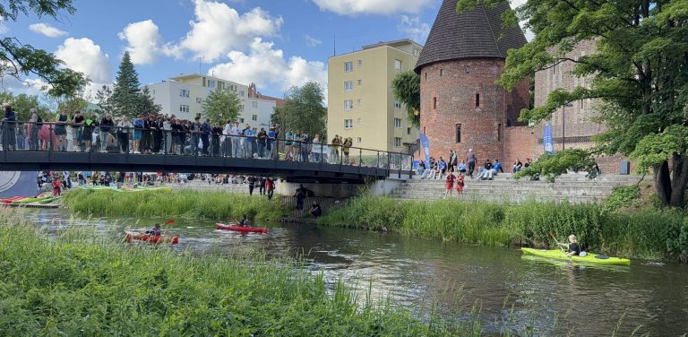 IV Międzyszkolne Regaty Słupia. Widok kajakarzy płynących rzeką Słupią i przepływających pod mostem, na którym stoją widzowie. W tle zabytkowa Baszta Czarownic