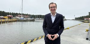 Zdjęcie przedstawia Jana Szyszko, Wiceministra Funduszy i Polityki Regionalnej, ubranego w elegancki, ciemny garnitur i białą koszulę, stojącego na nabrzeżu portu w Łebie. Uśmiecha się, patrząc prosto w obiektyw, a jego ręce są złożone w geście przypominającym kształt serca. Za nim rozciąga się widok na kanał portowy oraz marinę. Po lewej stronie, w oddali, widać zacumowane łodzie i jachty o różnych rozmiarach oraz budynki portowe. Po prawej stronie, w tle, można dostrzec białą latarnię morską oraz fragmenty zabudowań nadbrzeżnych. Nabrzeże, na którym stoi minister, jest betonowe, z charakterystycznymi żółto-czarnymi pasami ostrzegawczymi. Niebo jest jasne, chociaż zachmurzone