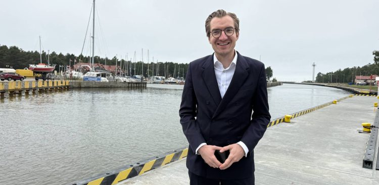 Zdjęcie przedstawia Jana Szyszko, Wiceministra Funduszy i Polityki Regionalnej, ubranego w elegancki, ciemny garnitur i białą koszulę, stojącego na nabrzeżu portu w Łebie. Uśmiecha się, patrząc prosto w obiektyw, a jego ręce są złożone w geście przypominającym kształt serca. Za nim rozciąga się widok na kanał portowy oraz marinę. Po lewej stronie, w oddali, widać zacumowane łodzie i jachty o różnych rozmiarach oraz budynki portowe. Po prawej stronie, w tle, można dostrzec białą latarnię morską oraz fragmenty zabudowań nadbrzeżnych. Nabrzeże, na którym stoi minister, jest betonowe, z charakterystycznymi żółto-czarnymi pasami ostrzegawczymi. Niebo jest jasne, chociaż zachmurzone
